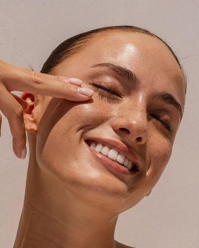 Smiling person gently touches their cheek with closed eyes under bright light.