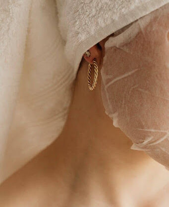 Woman with towel on head and facial sheet mask wearing gold hoop earring.